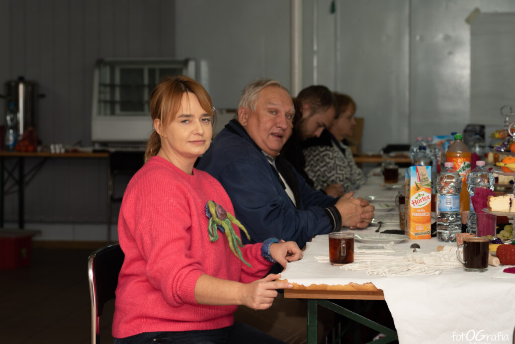 Zdjęcie przedstawia osoby siedzą ceprzy stole, na którym są ciasta, napoje.